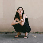 Woman in a black dress squatting against a plain wall.