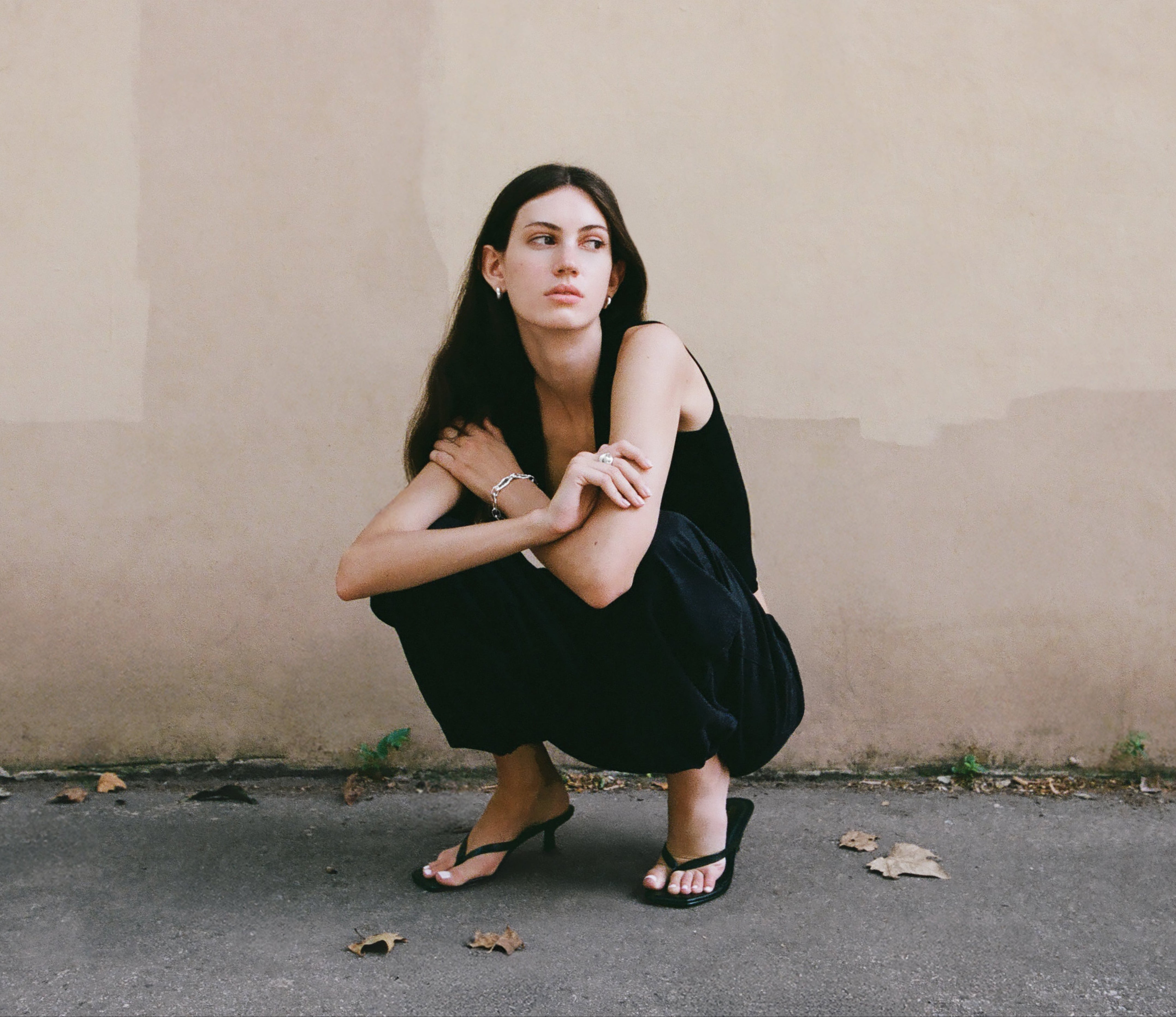 Woman in a black dress squatting against a plain wall.